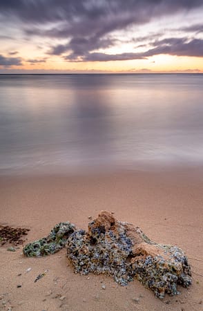 a rock on a sandy beach next to the ocean in Hawaii at sunset