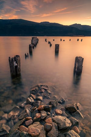 a sunrise over a body of water with rocks and abandoned dock in the foreground