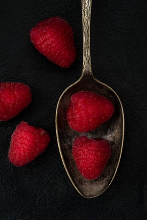 a still life of a spoon with raspberries on a black background
