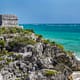 a mayan ruin on a rock near the ocean with Tulum in the background
